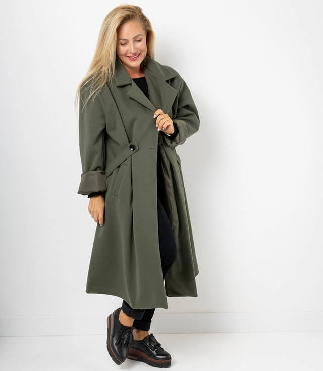 Frau in olivgrünem Trenchcoat mit tailliertem Schnitt, lässig posierend und lächelnd.