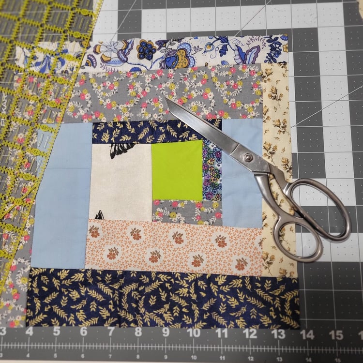 A log cabin block with a green center placed on a cutting mat. A pair of scissors and a ruler lie on top of it.