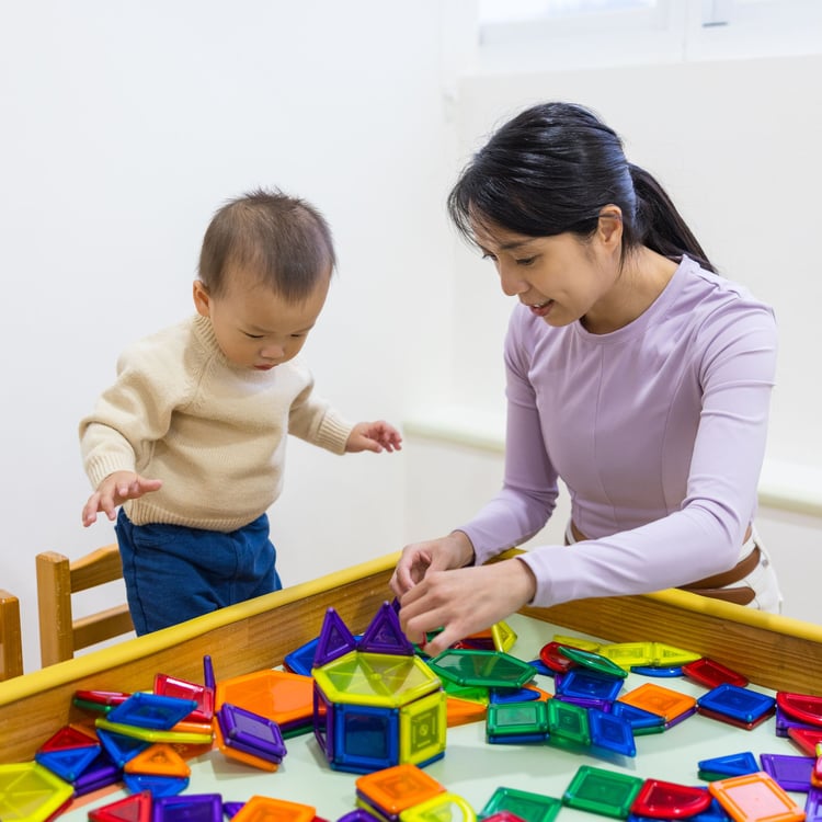 Mother playing bilateral coordination games with toddler at table, building shapes together to develop fine motor skills.