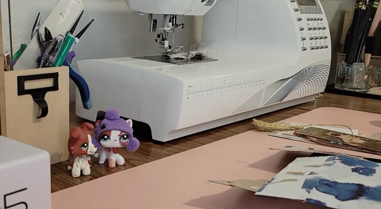 Two littlest pet shops sit on a desk in front of a sewing machine and cup of craft supplies
