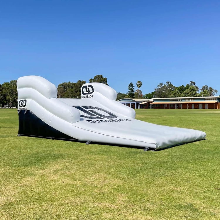 Mountain bike rider mid-air performing a jump above a black 20x13x5ft inflatable air bag landing pad at an outdoor bike park showing action sports safety equipment.