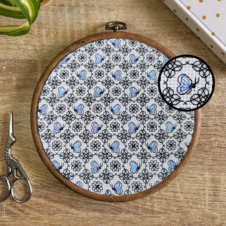 Modern blackwork embroidery pattern of small blue butterflies and black geometric flowers framed in a wooden embroidery hoop