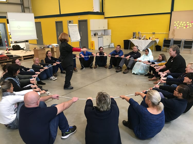 a team development consultant standing and facilitating from the centre of a circle of seated team members all holding a rope which is creating a perfect circle