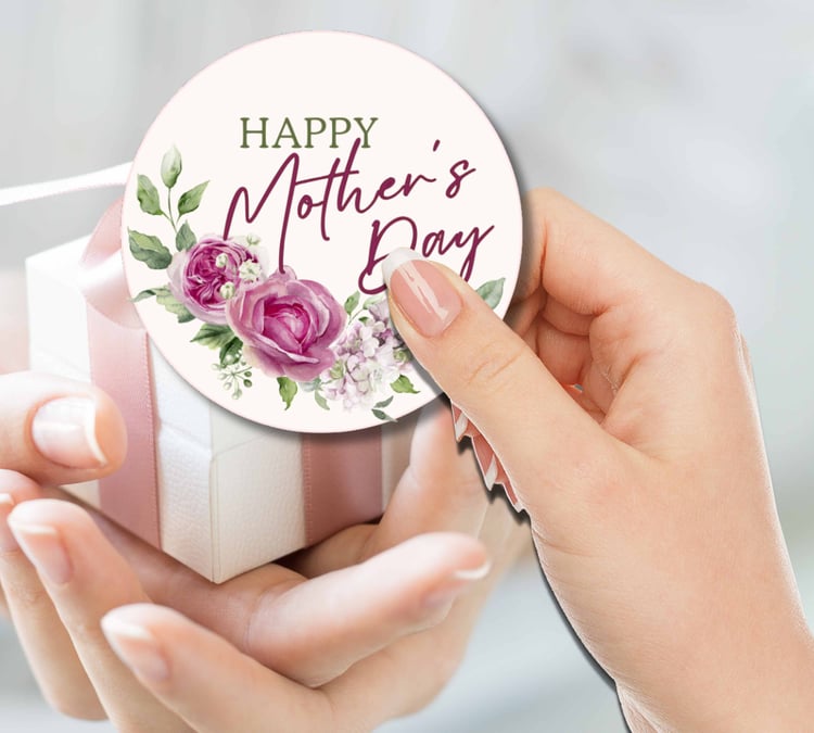 Hands holding a small white gift box with a pink ribbon, attaching a round floral card that reads “HAPPY Mother’s Day.
