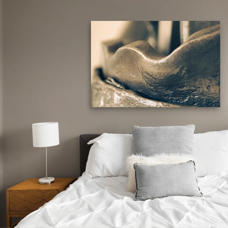 large horizontal canvas print of an abstract metallic curve hangs on a taupe wall above a bed with white bedding and gray pillows in a modern bedroom.