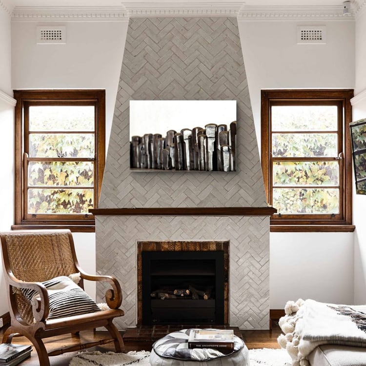 A large horizontal print of textured industrial metalwork hangs on a gray herringbone tiled fireplace in a bright, modern living room with a woven accent chair.