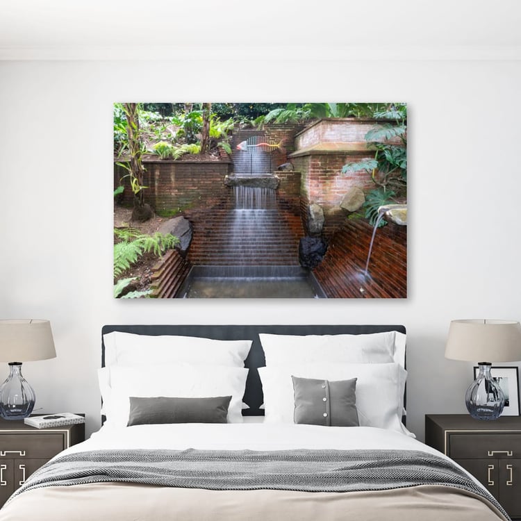 A large horizontal print of a tropical brick fountain hangs above a neatly made bed with white and gray linens in a bright, modern bedroom.