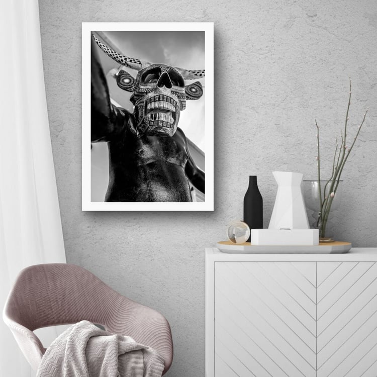 A vertically oriented matted white frame containing a black and white photograph of a masked Oaxacan performer hangs on a textured grey concrete wall above a white modern side cabinet and a soft pink lounge chair.