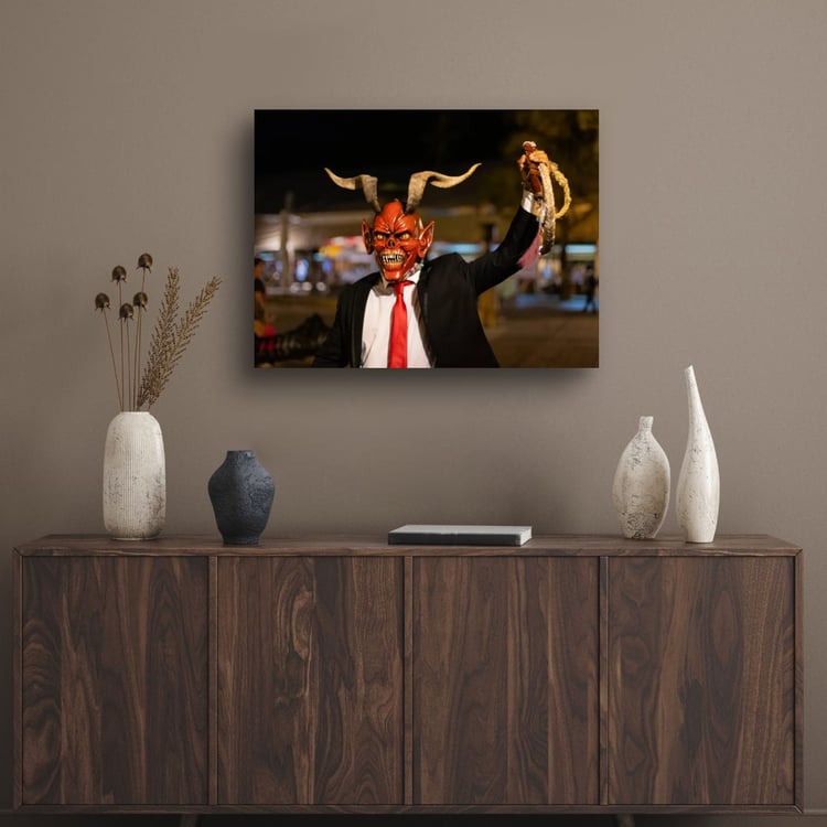 A horizontal gallery print of a red-masked devil character hangs on a muted brown wall centered above a dark walnut wood credenza styled with minimal ceramic vases.