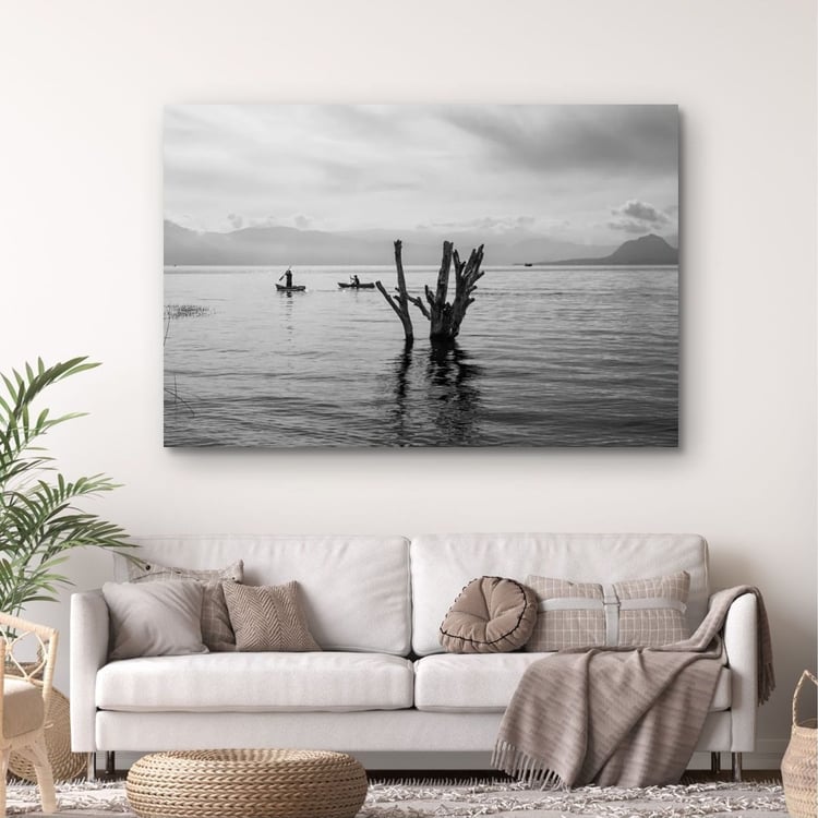 A large horizontal canvas print of a minimalist black and white lake photograph hangs on a white wall in a warm Japandi-style living room above a cream fabric sofa with neutral cushions.