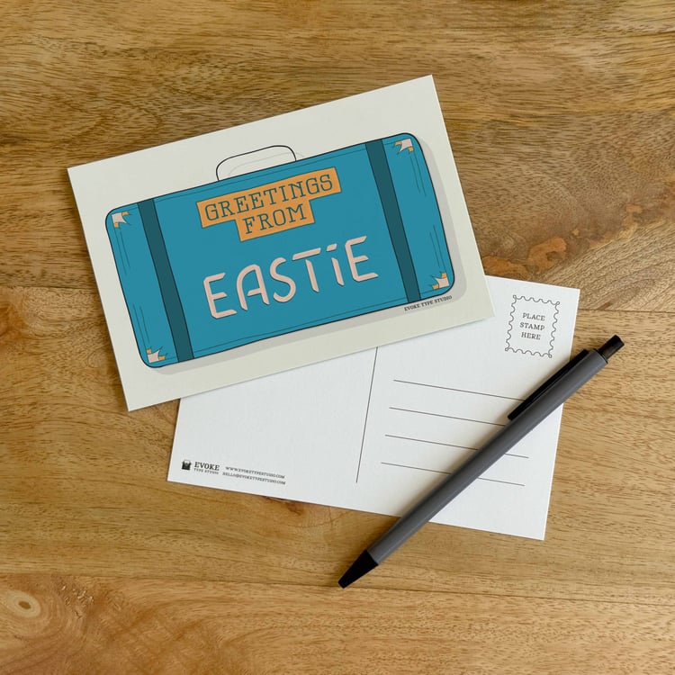 The front and back sides of a postcard with a pen placed on top. The front features a vintage suitcase illustration and the back shows the address lines and stamp indicator.