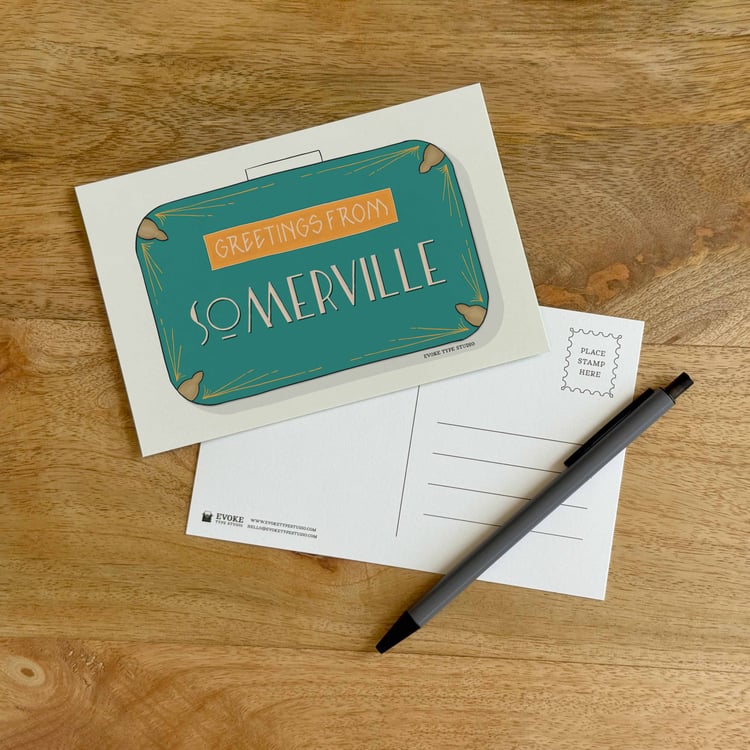 The front and back sides of a postcard with a pen placed on top. The front features a vintage suitcase illustration and the back shows the address lines and stamp indicator.