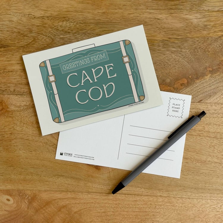 The front and back sides of a postcard with a pen placed on top. The front features a vintage suitcase illustration and the back shows the address lines and stamp indicator.