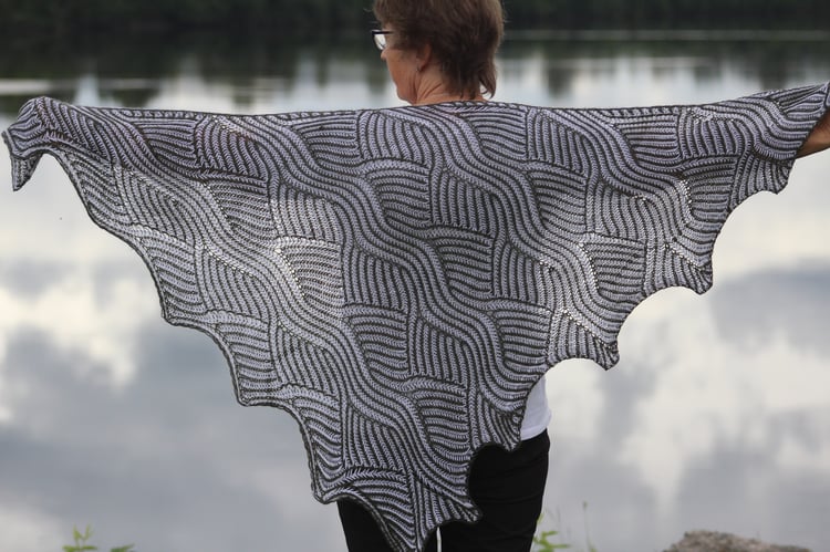 A grey and white Waterfall Shawl is shown in front of a body of water