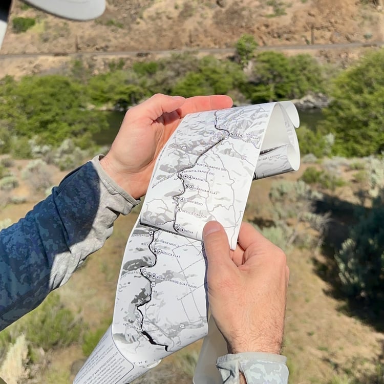 Angler holding the Lower Deschutes Ribbon Map open above the river canyon, showing campsites and rapids from Maupin downstream, with sagebrush hillsides and the Deschutes River visible below
