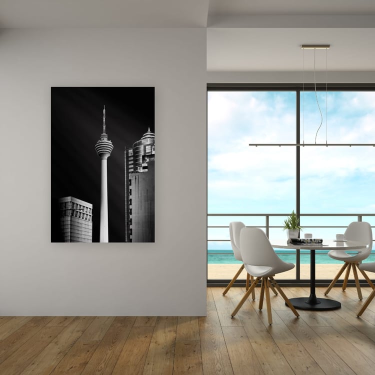 A large vertical frameless canvas print of a black and white Kuala Lumpur skyscraper scene hangs on a white dining room wall next to a large glass door overlooking the ocean.