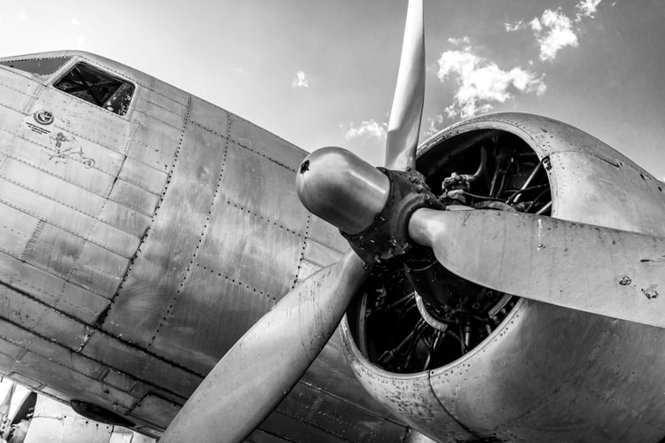 A dramatic, low-angle black and white photograph focusing closely on a vintage propeller and radial engine cowled on a metallic, riveted airplane fuselage under a sky with light clouds.