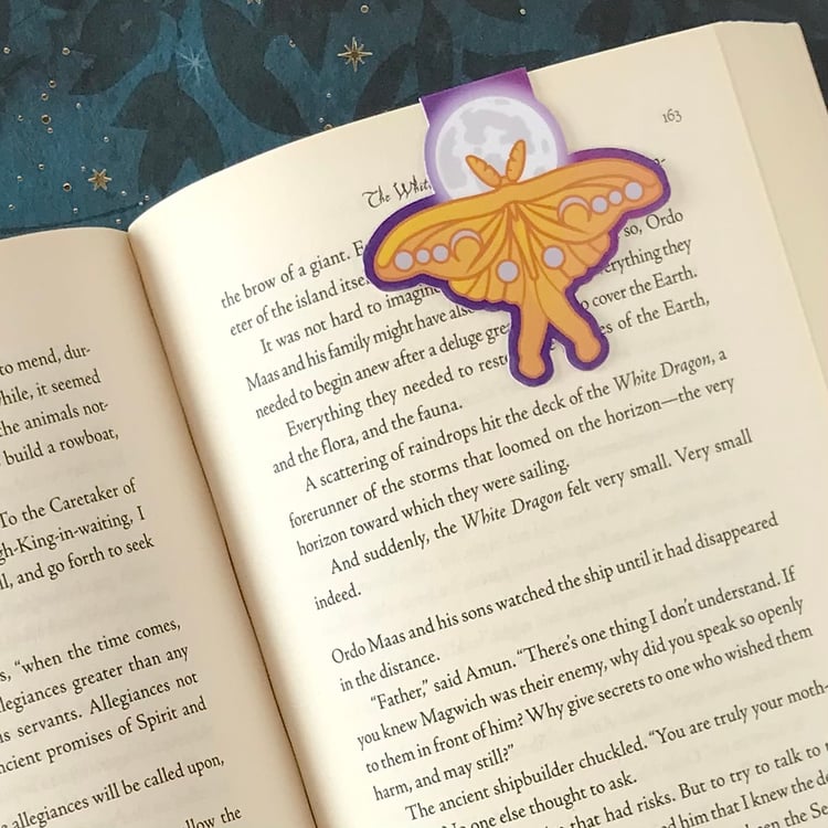 A luna moth- and moon-themed magnetic bookmark shown in use inside a fantasy novel.