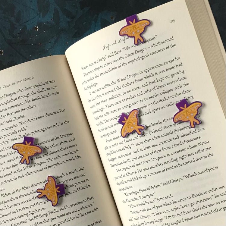 Five miniature magnetic bookmarks with a luna moth motif are shown in-use, marking a page in a fantasy novel.