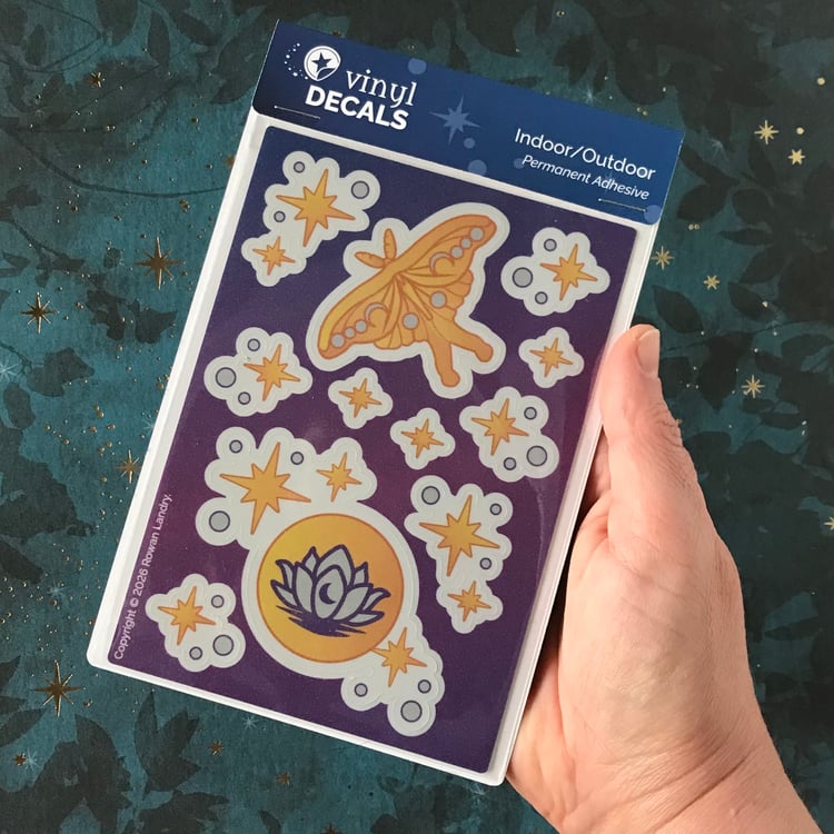 A sticker sheet featuring a luna moth and moon with lotus, plus stars, shown in-hand for scale.