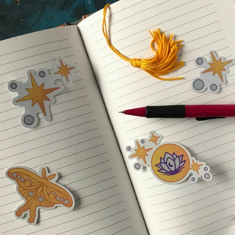 A set of four celestial-themed decals, featuring a luna moth, stars, moon and lotus sit upon a personal journal.