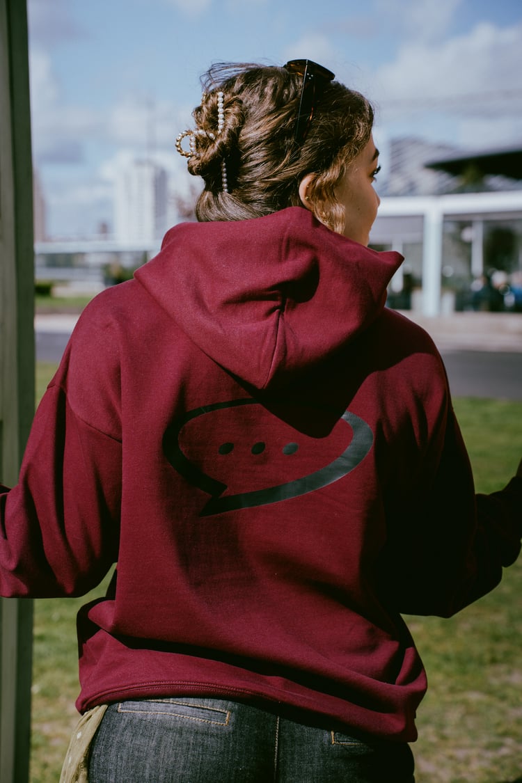 Foto van de achterkant van de Spraakuhloos Premium Hoodie in de kleur Wine. Het model heeft bruin opgestoken haar en staat met haar rug naar de camera. Op de achtergrond zie je een grasveld en een skyline.