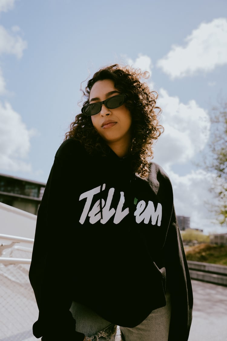 Foto van een vrouw met krullend zwart haar en een zwarte zonnebril. Ze staat enigszins voorover gebogen en kijkt direct en zelfverzekerd in de camera. Ze draagt de Tell 'em Basic Hoodie in de kleur Black met een groot wit Tell 'em logo op de borst. Op de 