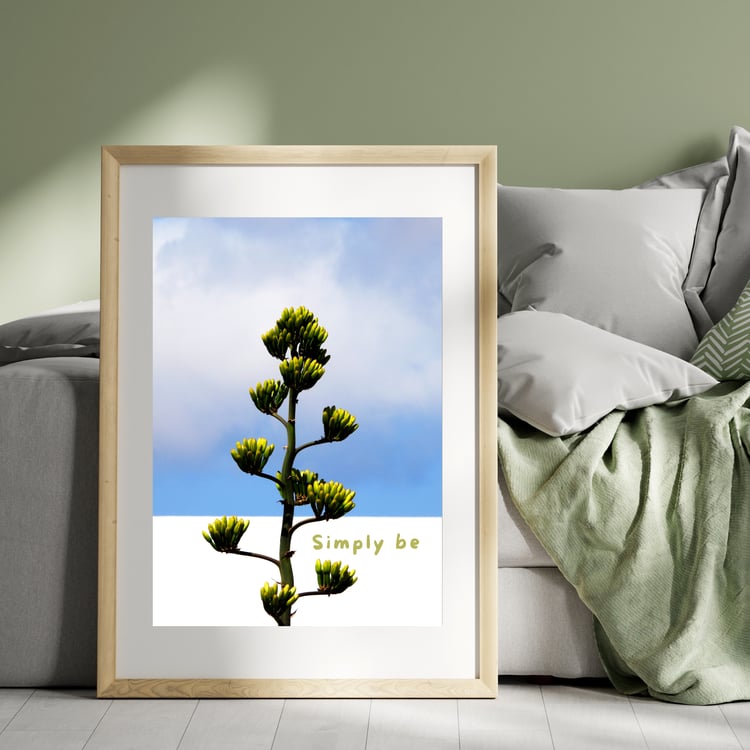 A minimalist wooden frame mockup resting on the floor next to a cozy grey sofa with cushions and a sage green blanket. The framed print features an original nature photograph of a green agave plant against a blue sky with the text Simply be...