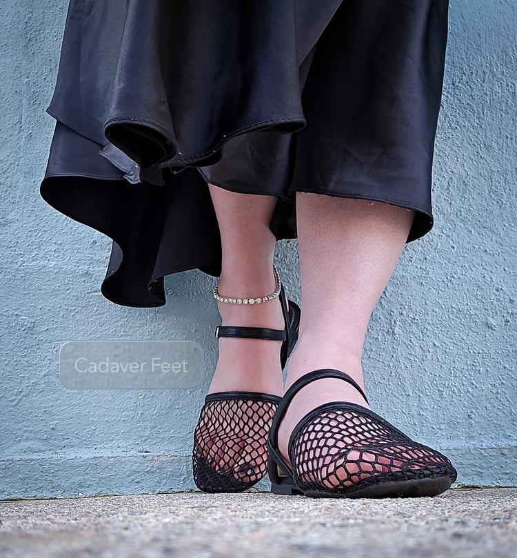the pastel blue of the iconic rainbow row house is a backdrop to a woman resting against the wall. long folds of black fabric from her skirt drape around her, as a diamond anklet adorns one delicate ankle. black strappy mesh sandals adorn her feet allowin