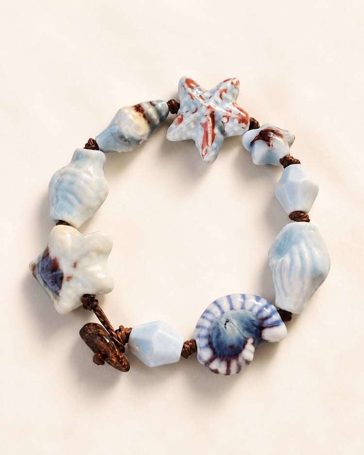 Dettaglio del bracciale artigianale con stella marina e conchiglia in ceramica smaltata, cordoncino marrone e componenti in acciaio inox.