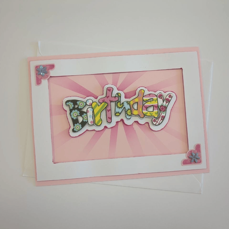 A landscape card in pink with a white frame. The word "Birthday" in multiple colours and patterns of bubble text floats over a pink blended starburst effect. Two small iridescent corner stickers sit in opposite corners of the frame.