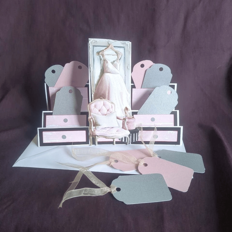 A stepper card with lower sides and a higher middle. The middle panel features an image of a pink chiffon prom dress hanging on a door, in front of which are a padded armchair and hat boxes. The steps of the card have been made to resemble drawers, and gi