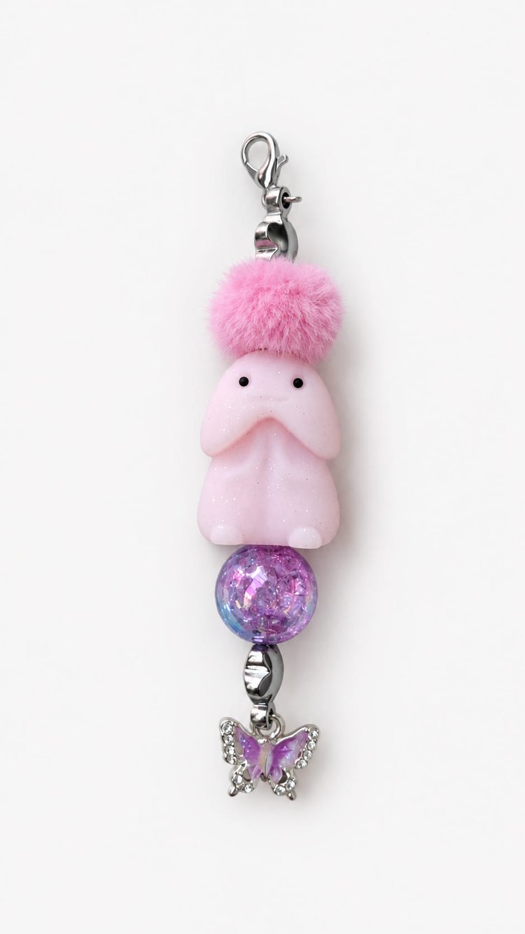 Detailed close-up of a pink character keychain featuring a fluffy pink pom-pom, a translucent purple crackle-glass bead, and a high-shine silver butterfly charm embedded with clear rhinestones.