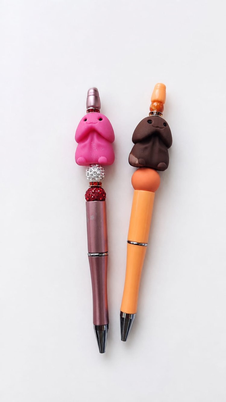 Two custom handmade novelty ballpoint pens standing side-by-side, displaying detailed stacked beads including pink and brown adult-themed focal charms, crystal spacer rings, and sparkling glitter beads.