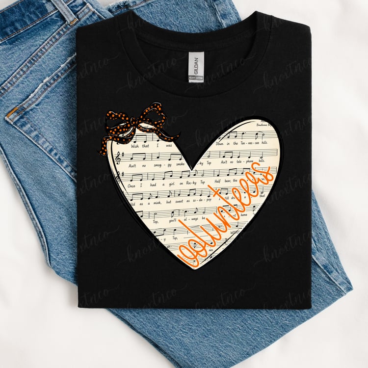 Black T-shirt featuring a heart-shaped vintage sheet music design with orange “Vols” script lettering and an orange-and-black polka dot bow, styled with blue denim jeans.