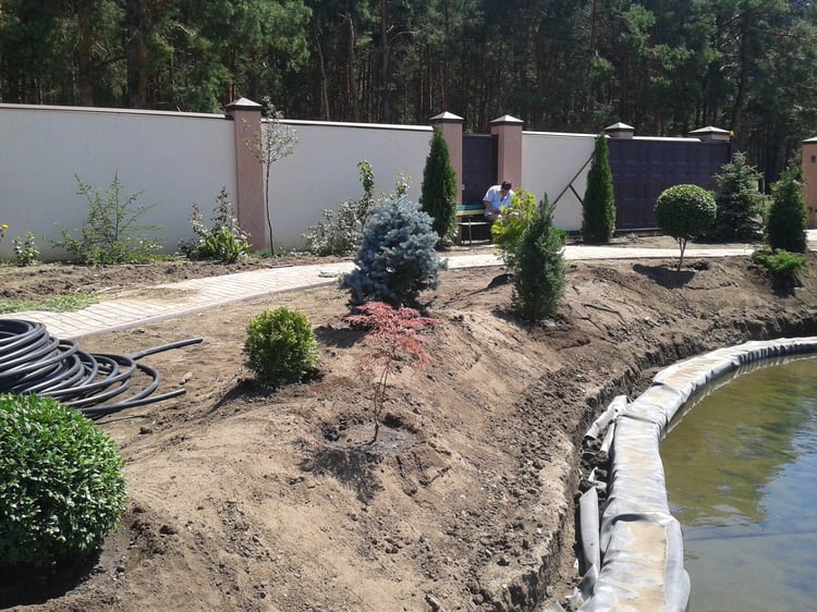 Natural swimming pond construction showing shoreline landform modelling, slope stabilisation, stone finishing and preparation of planted earth mounds in a large private garden.