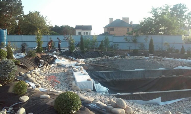 Natural swimming pond construction showing shoreline landform modelling, slope stabilisation, stone finishing and preparation of planted earth mounds in a large private garden.