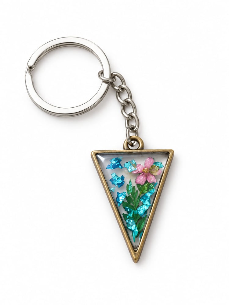 Handmade triangle botanical resin keychain featuring a real pressed pink five-petal flower with green leaves and vibrant metallic blue foil flakes suspended in clear resin inside a vintage bronze frame.
