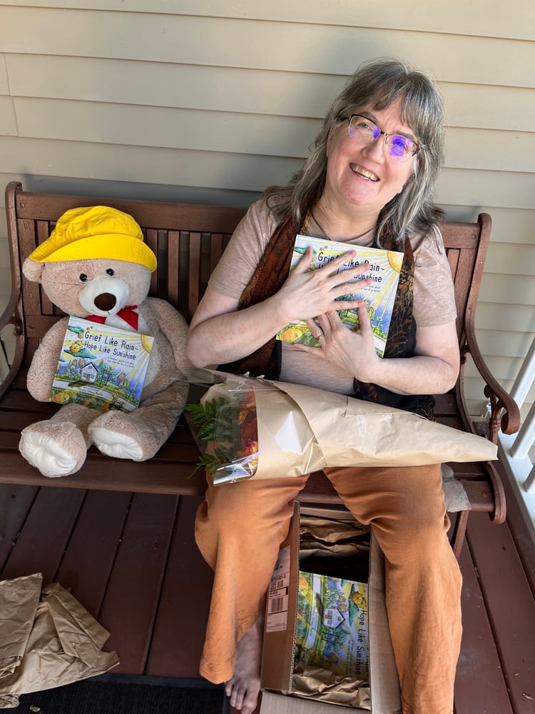 Patricia Tiffany Morris holding Grief Like Rain—Hope Like Sunshine for the first time, stuffed bear also holding a book. Box opened on porch.