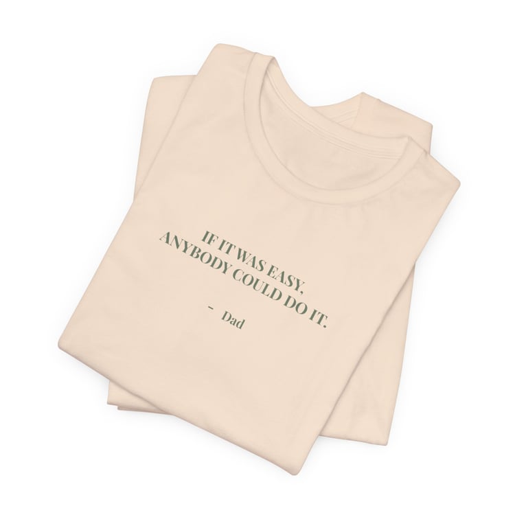 Folded cream t‑shirt with the quote ‘If it was easy, anybody could do it. — Dad’ printed on the front.”