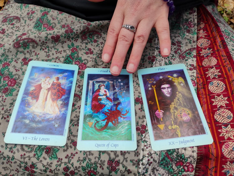 Photograph of Tarot reader Evylyn Rose (only her left hand is visible) pointing to a 3-card spread featuring Major Arcana cards (The Lovers & Death) and Minor Arcana cards (Queen of Cups). Photograph by Ryan Hobbs, 2025.