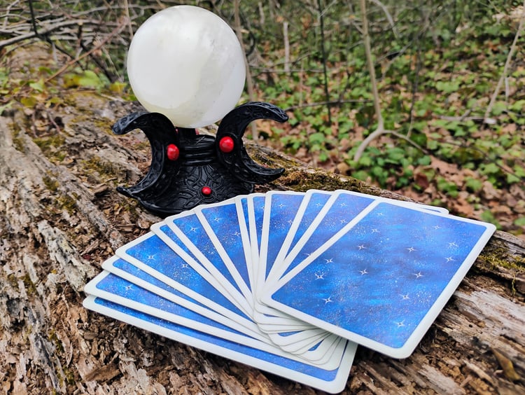 photograph of fanned out celestial Tarot cards on a log in front of a selenite cyrstal ball resting in a black and red crescent moon holder; photograph taken by Ryan Hobbs, 2025