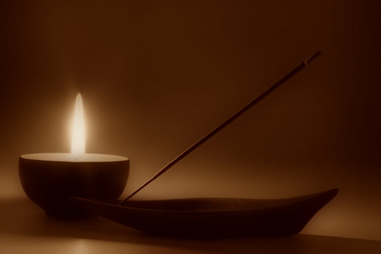 stock photo with simple tan-brown background depicting a small candle and incense holder with lit incense stick