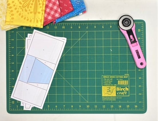 A quilter has prepared tools for paper piecing, a cutting mat, rotary cutter, printed foundation pattern and fabrics.