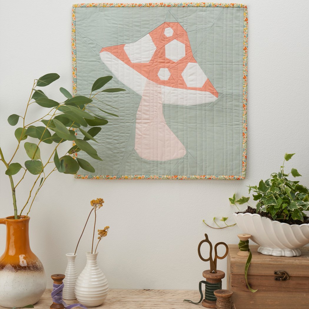 A cute coral and white mushroom mini quilt with a pastel green background hanging on a wall behind some rustic wooden sewing spools and foliage