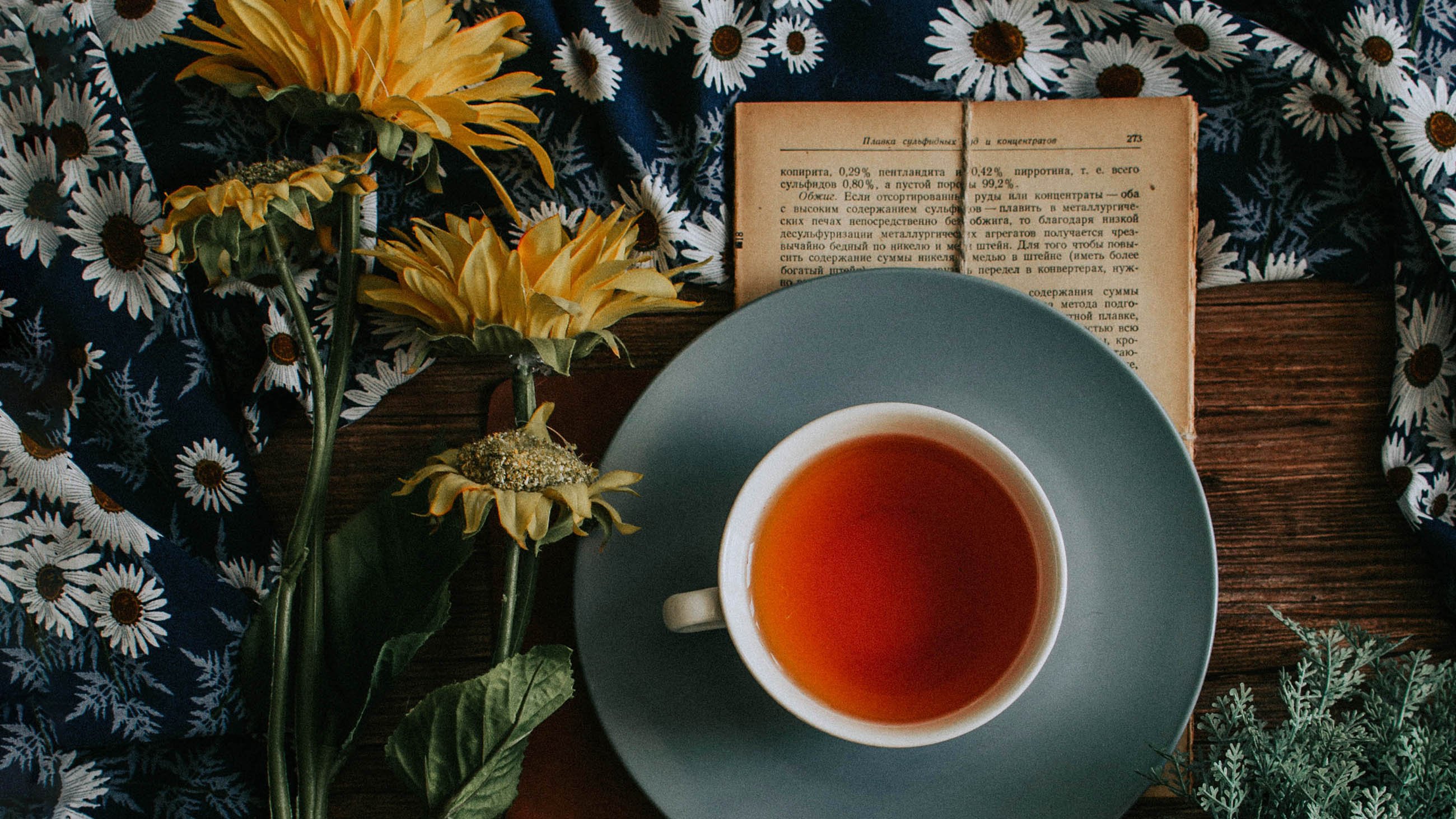 Cozy still of a cup of tea sitting on an open book next to some sunflowers and floral fabric on a wooden table.