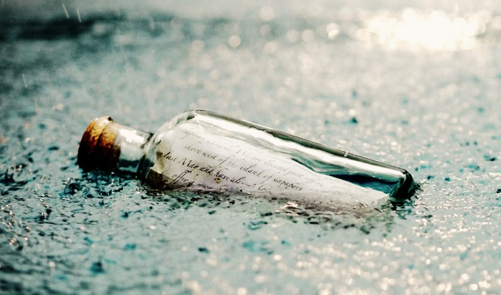 A clear glass bottle with a brown cork resting in shallow, rain-speckled water near a shore. Inside the bottle is an elegantly handwritten letter on white paper, sealed with a vintage feather quill script style. Soft moonlight glows gently on the bottle, evoking a mysterious, sacred message waiting to be discovered - inviting viewers to unlock hidden truths and ancient wisdom