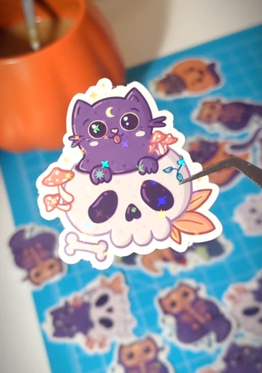 A sticker of a purple cat sitting on top of a skull. The cat has a cute expression with sparkling eyes and a crescent moon on its forehead. It is surrounded by mushrooms and leaves, with a bone at the bottom. The design features soft pastel colors and small sparkling details, giving it a playful, whimsical Halloween vibe. In the background, more Halloween-themed stickers are visible.