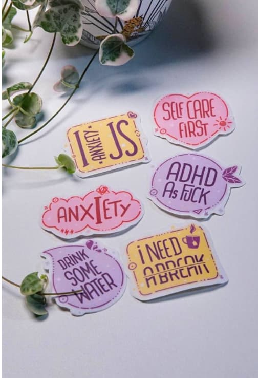 A collection of motivational and self-care stickers laid out around a plant. The stickers include phrases like "I Need A Break," "Self Care First," "Drink Some Water," "ADHD As F*ck," and "Anxiety Is." Each sticker has a vibrant color palette, with shades of pink, purple, and yellow