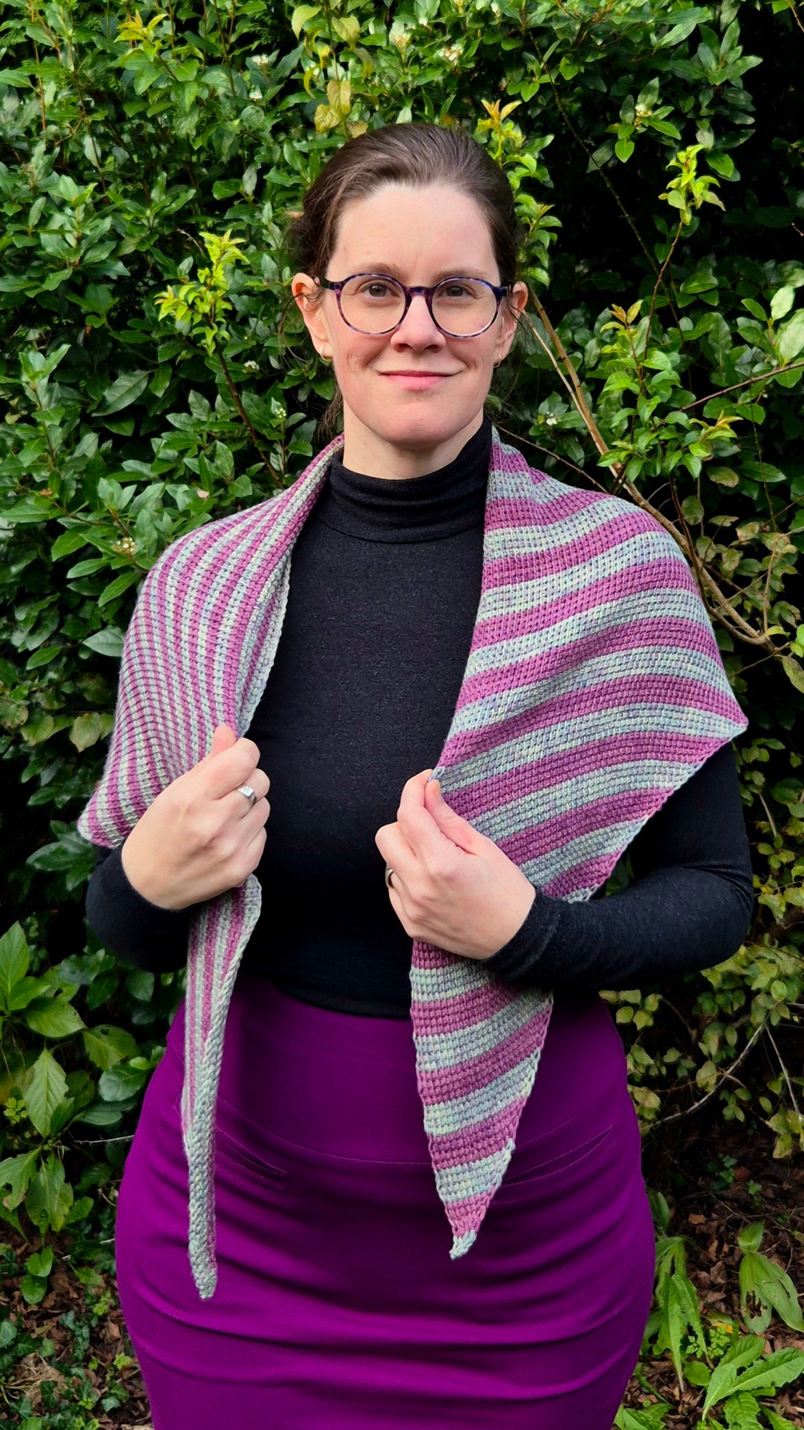 A Tunisian crocheted shawl in purple and green stripes that are vertical on the left side and horizontal on the right side. The shawl is a symmetrical triangle, worn across the back, but the image shows the two outer points draped over the shoulders in th